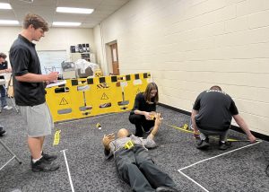 three students work on a mock crime scene with a dummy body laying on the ground