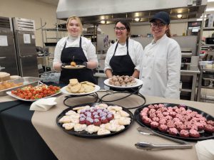 three culinary students stand in a kitchen with platters of food in front of them