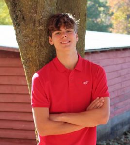 a boy with his arms crossed and in a red shirt leans up against a tree with a red barn in the background