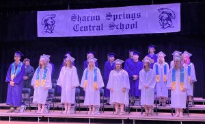 graduates stand in gowns and caps under a sign that says Sharon Springs Central School