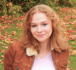 a smiling girl with a brown jacket and fall leaves in the background