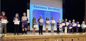 young students stand in a line on stage holding certificates with a screen behind them that says summer reading