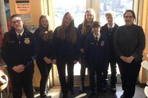 Seven students stand in a row in front of a window wearing FFA jackets.