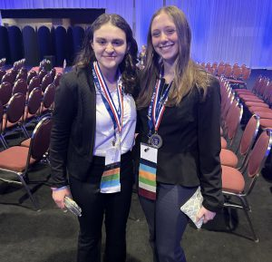 Two students stand in an auditorium with medals around their necks.