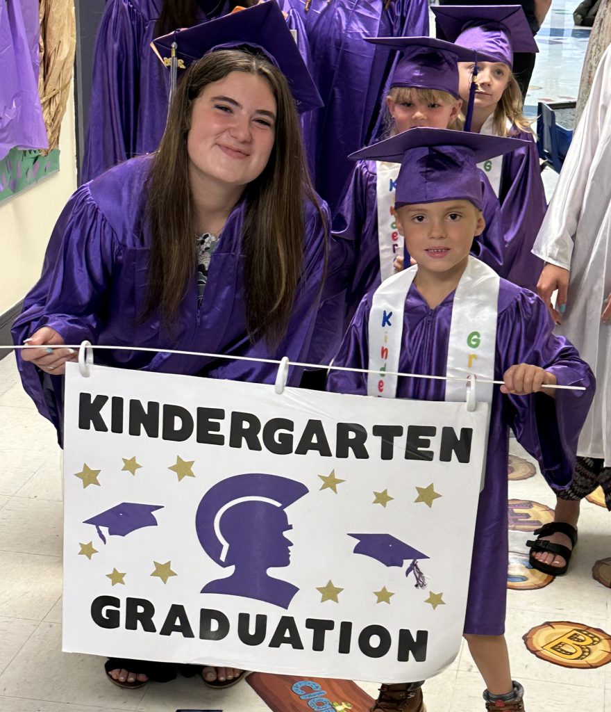 girl and boy in cap and gown carrying kindergarten banner